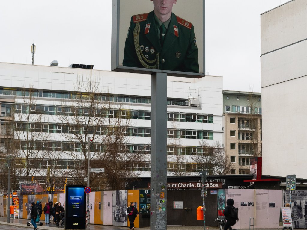 Checkpoint Charlie on a rainy day. In the center of the photo is the lightbox rising above the street. It shows a larger than scale photo of a russian soldier. In the background is an intersection and the Checkpoint Charlie BlackBox.