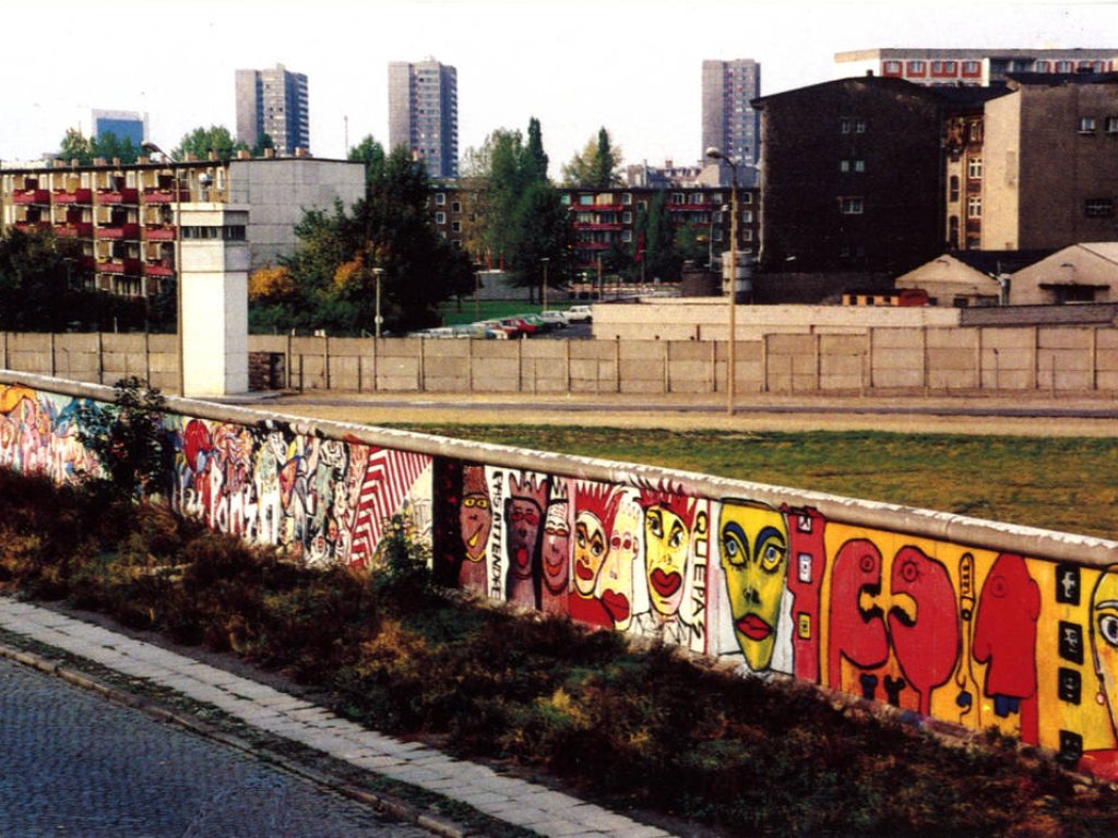 Im unteren Bildteil ist die bunt bemalte Mauer zu sehen. Darüber sieht man in den Grenzstreifen, wo ein Wachturm steht.