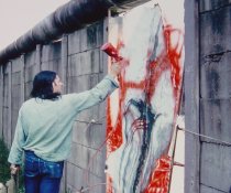 Elsner steht auf einem Haufen herausgebrochener Mauerzeile. Neben ihm in der Mauerlücke ist sein abstraktes Bild eingesetzt, das er mit einer Sprühpistole mit roter Farbe weiter bearbeitet.