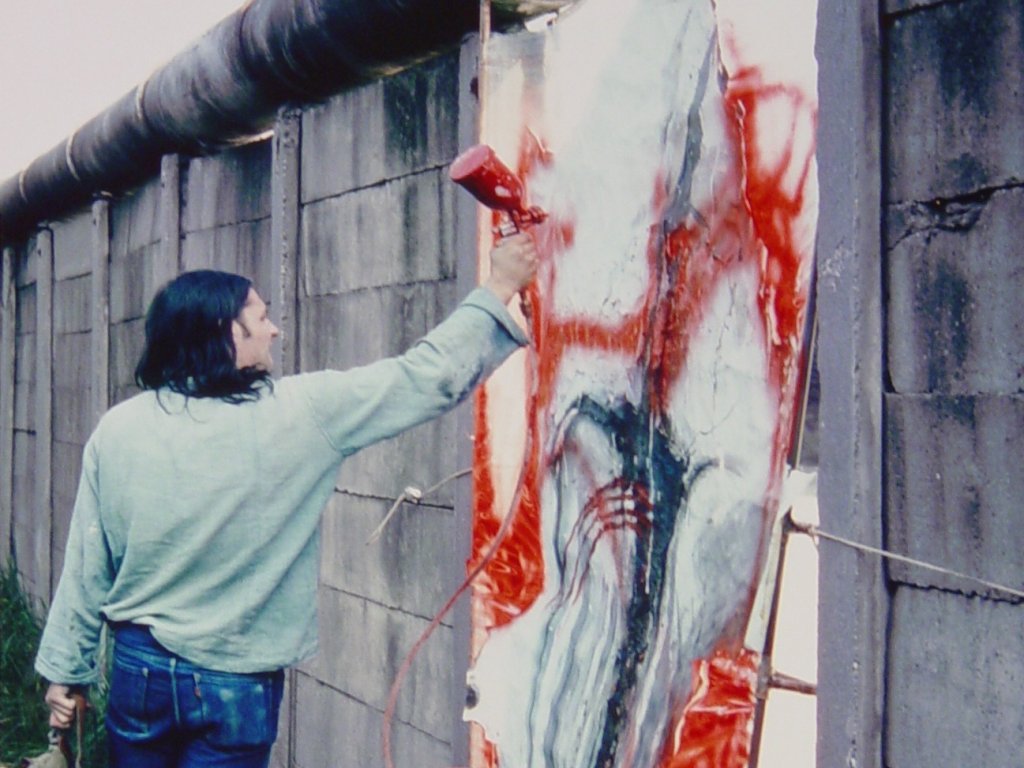 Elsner steht auf einem Haufen herausgebrochener Mauerzeile. Neben ihm in der Mauerlücke ist sein abstraktes Bild eingesetzt, das er mit einer Sprühpistole mit roter Farbe weiter bearbeitet.