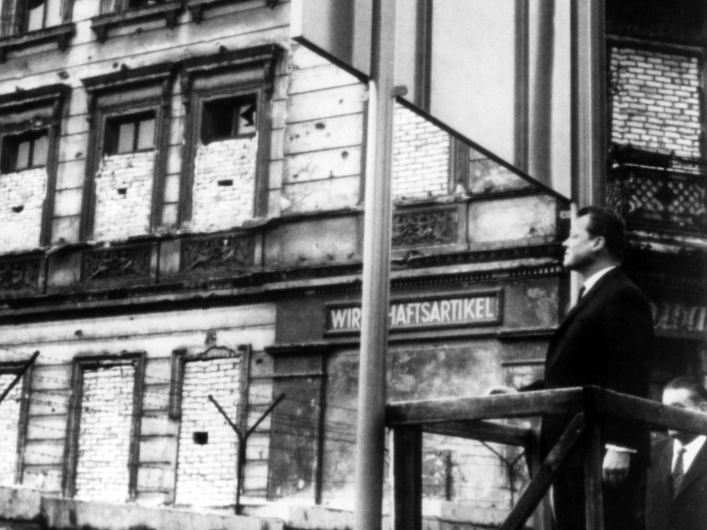 1964: West-Berlins Regierender Bürgermeister Willy Brandt steht am dritten Jahrestag des Mauerbaus auf einem Aussichts-Podest in der Bernauer Straße und blickt auf den Ostteil der Stadt. Die Bernauer Straße hat Symbolcharakter für die Teilung der Stadt, unter anderem weil hier am 22. August 1961 die erste Mauertote, Ida Siekmann, ums Leben gekommen ist. Willy Brandt wird es in seiner späteren Rolle als Bundeskanzler mit seiner „Neuen Ostpolitik“ und dem Credo „Wandel durch Annäherung“ gelingen, die starre Konfrontation der beiden deutschen Staaten zu lösen.
