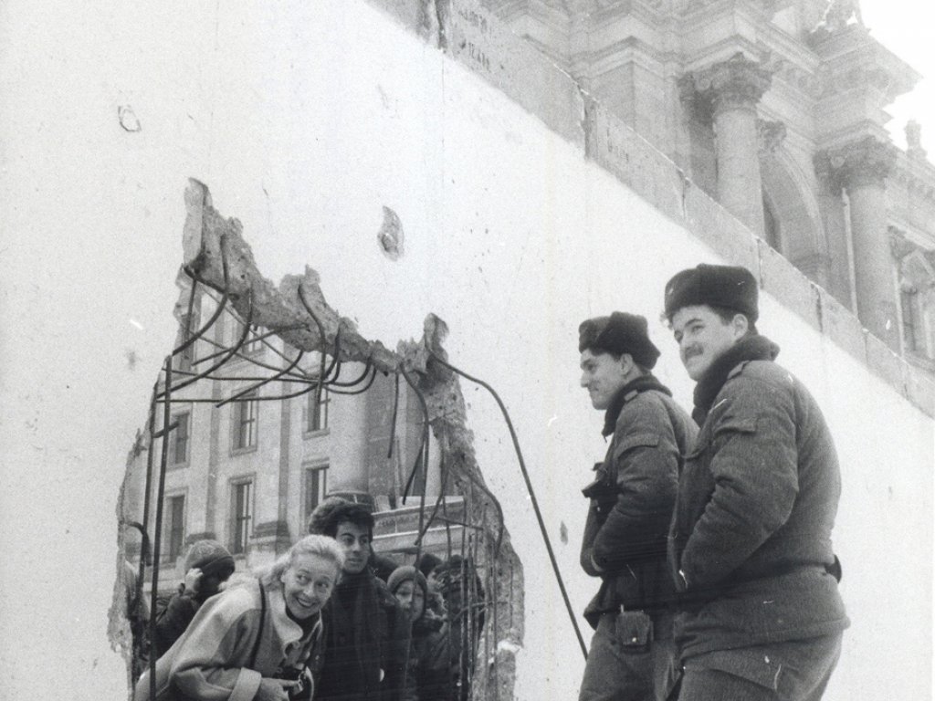 Mauerdurchbruch am Berliner Reichstagsgebäude, Januar 1990 Eine Frau steckt grinsend den Kopf durch ein großes Loch in der Mauer, weitere Menschen stehen hinter ihr. Auf der anderen Seite im Vordergrund stehen zwei lächelnde Grenzsoldaten, die sich mit den Menschen durch das Loch zu unterhalten scheinen. Im Hintergrund ist das Reichstagsgebäude zu erkennen.