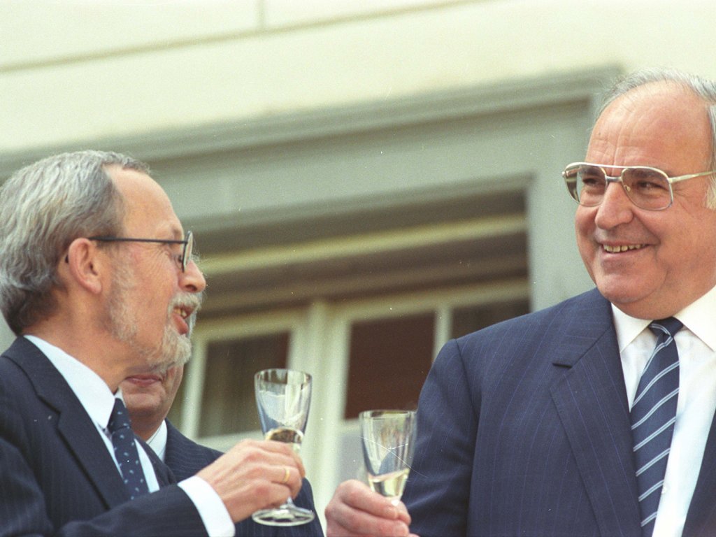 Lothar de Maizière und Helmut Kohl stehen im Freien vor einem Fenster. Sie halten jeweils ein Glas Sekt in der Hand, schauen sich lächelnd an und prosten sich zu.