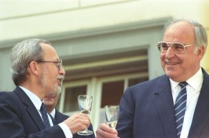Lothar de Maizière und Helmut Kohl stehen im Freien vor einem Fenster. Sie halten jeweils ein Glas Sekt in der Hand, schauen sich lächelnd an und prosten sich zu.