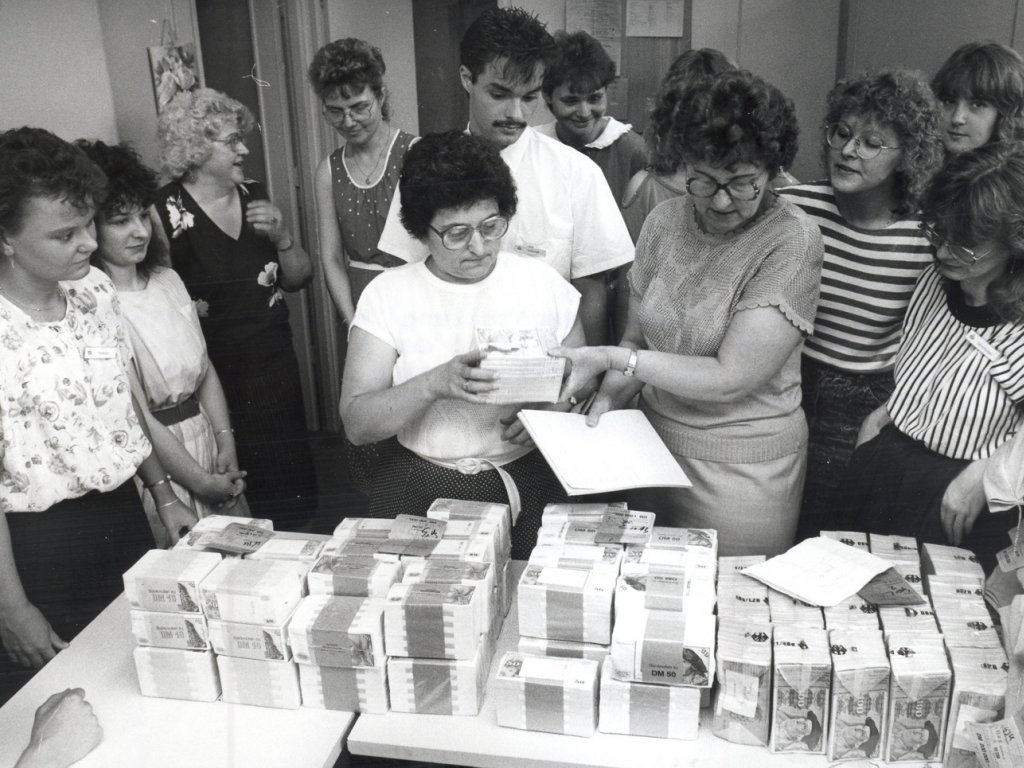 Auf einem Tisch liegen dutzende Bündel mit Hundertmarkscheinen. Um den Tisch stehen Leute, unterhalten sich und blicken auf das Geld. Eine Frau hält in einer Hand ein Blatt Papier und reicht der Frau neben sich mit der anderen Hand ein Geldbündel.