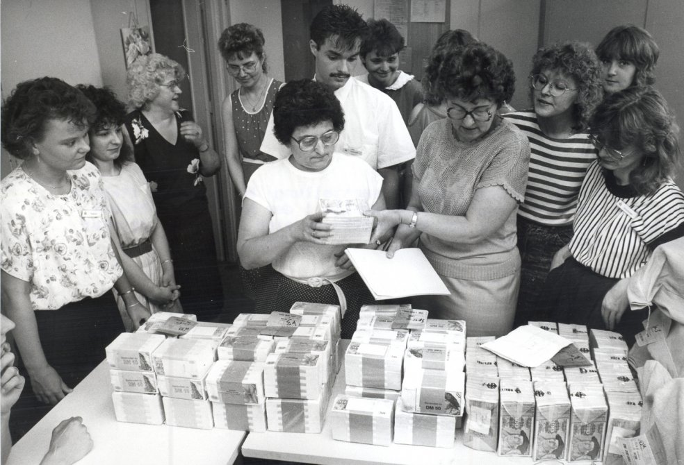 Auf einem Tisch liegen dutzende B&uuml;ndel mit Hundertmarkscheinen. Um den Tisch stehen Leute, unterhalten sich und blicken auf das Geld. Eine Frau h&auml;lt in einer Hand ein Blatt Papier und reicht der Frau neben sich mit der anderen Hand ein Geldb&uuml;ndel.