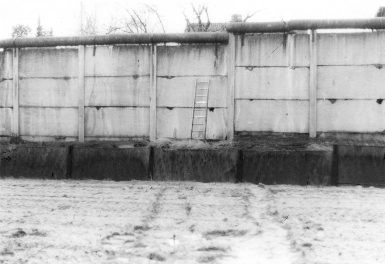 Das Foto ist innerhalb des Grenzstreifens aufgenommen. Zentral im Bild steht eine Leiter an die Mauer gelehnt.