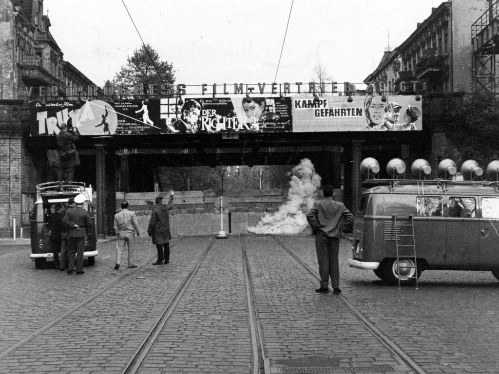 Im Bildzentrum ist eine Bahnbrücke, die darunter verlaufende Straße ist zugemauert. Links steht ein Transporter, daneben ein paar Männer. Auf dem Dach des Transporters filmt ein Kameramann Richtung Mauer, vor der eine Rauchschwade aufsteigt. Rechts im Vordergrund steht ein Wagen des Studios am Stacheldraht, die sechs Lautsprecher auf dem Dach sind auf die Mauer gerichtet.