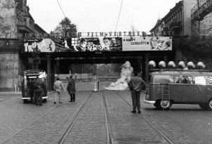 Im Bildzentrum ist eine Bahnbrücke, die darunter verlaufende Straße ist zugemauert. Links steht ein Transporter, daneben ein paar Männer. Auf dem Dach des Transporters filmt ein Kameramann Richtung Mauer, vor der eine Rauchschwade aufsteigt. Rechts im Vordergrund steht ein Wagen des Studios am Stacheldraht, die sechs Lautsprecher auf dem Dach sind auf die Mauer gerichtet.