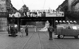 Im Bildzentrum ist eine Bahnbrücke, die darunter verlaufende Straße ist zugemauert. Links steht ein Transporter, daneben ein paar Männer. Auf dem Dach des Transporters filmt ein Kameramann Richtung Mauer, vor der eine Rauchschwade aufsteigt. Rechts im Vordergrund steht ein Wagen des Studios am Stacheldraht, die sechs Lautsprecher auf dem Dach sind auf die Mauer gerichtet.