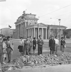 Eine Gruppe ratlos blickender Menschen steht auf der Straße. Vor ihnen liegen Steinhaufen, davor ist ein Graben. Im Hintergrund erhebt sich das Brandenburger Tor.