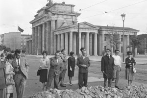 Eine Gruppe ratlos blickender Menschen steht auf der Straße. Vor ihnen liegen Steinhaufen, davor ist ein Graben. Im Hintergrund erhebt sich das Brandenburger Tor.