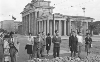 Eine Gruppe ratlos blickender Menschen steht auf der Straße. Vor ihnen liegen Steinhaufen, davor ist ein Graben. Im Hintergrund erhebt sich das Brandenburger Tor.