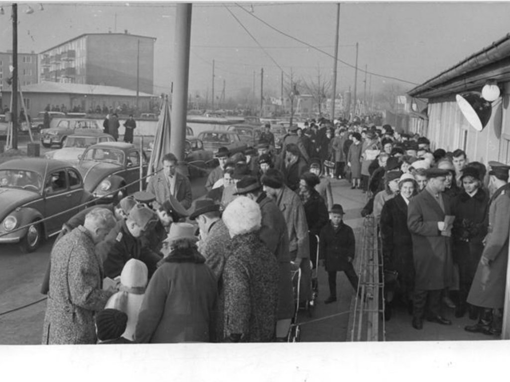 Im Bildzentrum ist eine große Ansammlung von Menschen, die Schlange stehen. Uniformierte Männer kontrollieren ihre Papiere. Rechts im Bild ist das Gebäude der Übergangsstelle, links eine Autokolonne.