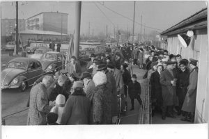 Im Bildzentrum ist eine große Ansammlung von Menschen, die Schlange stehen. Uniformierte Männer kontrollieren ihre Papiere. Rechts im Bild ist das Gebäude der Übergangsstelle, links eine Autokolonne.