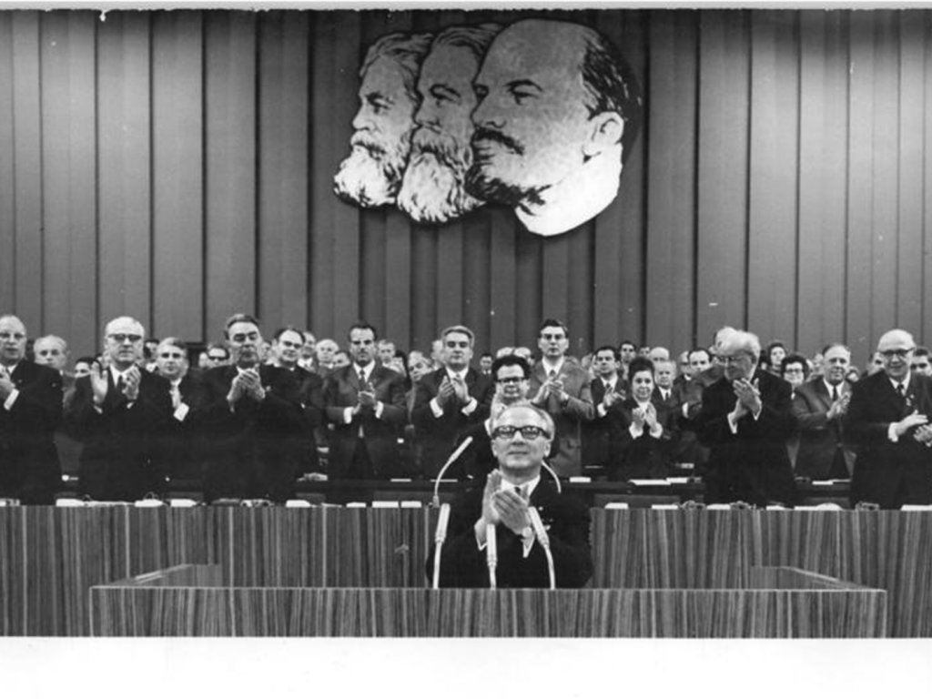 Erich Honecker kurz nach seinem Amtsantritt als Erster Sekretär des Zentralkomitees (ZK) auf dem VIII. Parteitag in Ost-Berlin. Hinter Leonid Breshnew (3. v. li.), Aufnahme 15. Juni 1971 Erich Honecker sitzt vor Mikrofonen und klatscht. Hinter ihm klatschen dutzende Männer. An der Wand über ihnen hängen die überlebensgroßen Konterfeis von Marx, Engels und Lenin.