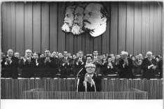 Erich Honecker sitzt vor Mikrofonen und klatscht. Hinter ihm klatschen dutzende Männer. An der Wand über ihnen hängen die überlebensgroßen Konterfeis von Marx, Engels und Lenin.