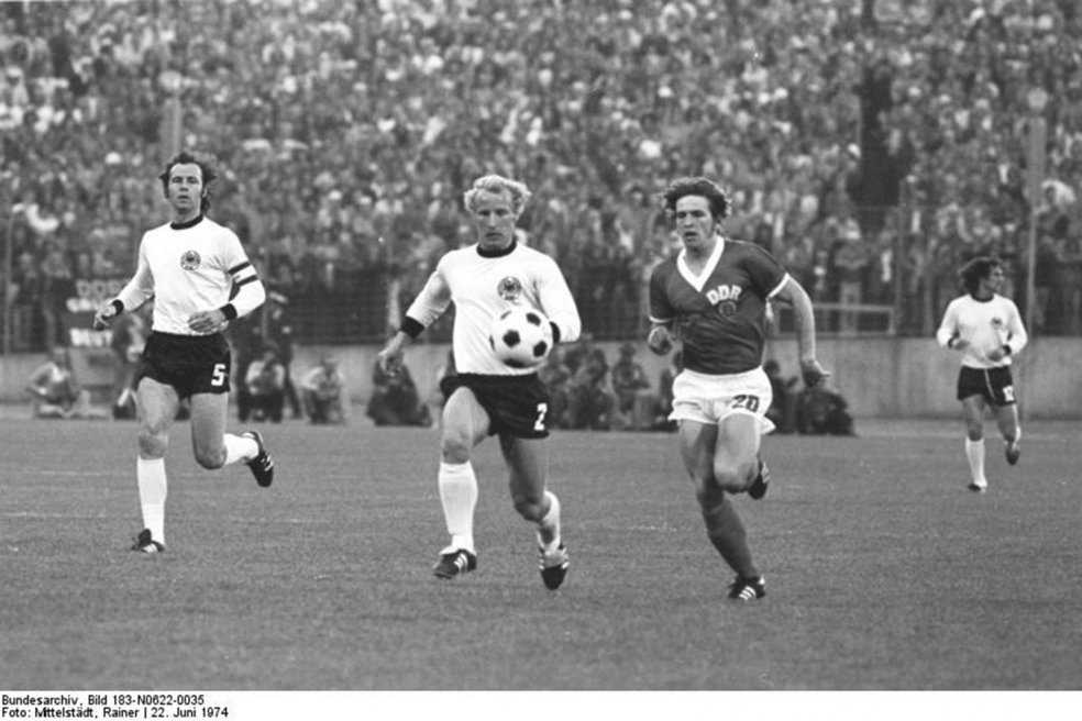 In der Mitte jagen Vogts und Hoffmann den Ball, links läuft Beckenbauer, rechts im Hintergrund ein weiterer Spieler der Bunderepublik. Im Bildhintergrund sind unscharf eine Reihe Fotografen und die vollbesetzte Zuschauertribüne zu erkennen.
