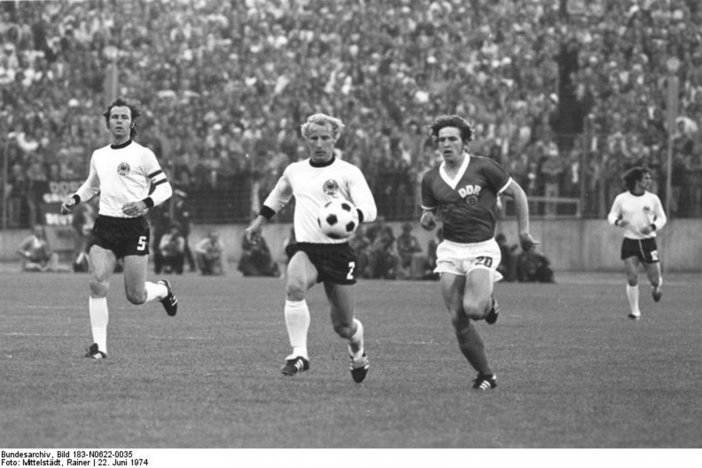 In der Mitte jagen Vogts und Hoffmann den Ball, links läuft Beckenbauer, rechts im Hintergrund ein weiterer Spieler der Bunderepublik. Im Bildhintergrund sind unscharf eine Reihe Fotografen und die vollbesetzte Zuschauertribüne zu erkennen.