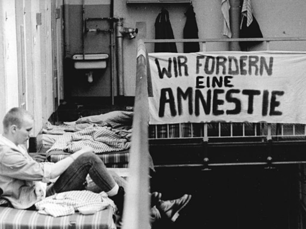 Die Insassen der Haftanstalten in der DDR (hier in Dresden) protestieren und fordern eine Generalamnestie; Aufnahme 28. September 1990  Ein Mann mit kurzgeschorenem Haar sitzt auf einer Matratze im Flur vor einer Zellentür. Am Geländer neben ihm hängt ein Transparent mit der Aufschrift: Wir fordern eine Amnestie.