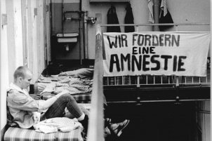 Ein Mann mit kurzgeschorenem Haar sitzt auf einer Matratze im Flur vor einer Zellentür. Am Geländer neben ihm hängt ein Transparent mit der Aufschrift: Wir fordern eine Amnestie.