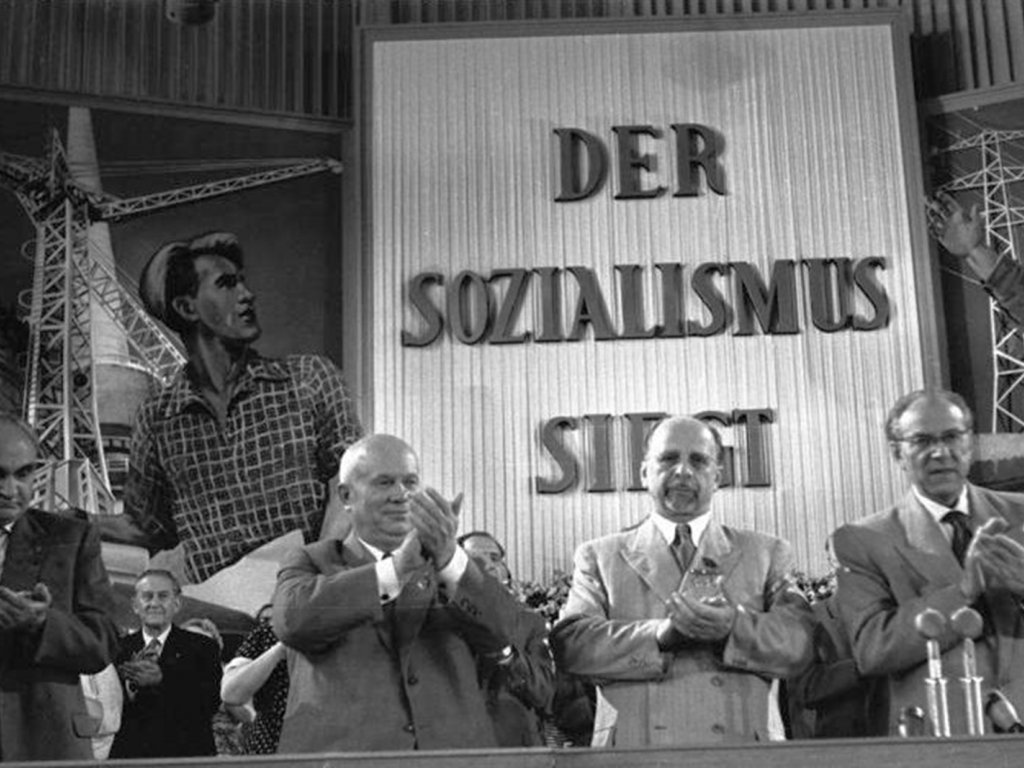 V. Parteitag der SED in der Ost-Berliner Werner-Seelenbinder-Halle (v. l. n. r.: Heinrich Rau, N.S. Chruschtschow, Walter Ulbricht und Otto Grotewohl); Aufnahme 10. Juli 1958 Die vier Politiker stehen klatschend in einer Reihe. Hinter ihnen sind weitere klatschende Menschen zu sehen und der riesige Schriftzug an der Wand: Der Sozialismus siegt.
