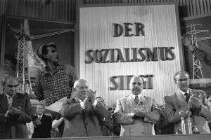 Die vier Politiker stehen klatschend in einer Reihe. Hinter ihnen sind weitere klatschende Menschen zu sehen und der riesige Schriftzug an der Wand: Der Sozialismus siegt.