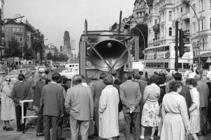 In der Bildmitte läutet die riesige Glocke, mit dem Rücken zur Kamera stehen Schaulustige davor. Im Bildhintergrund steht die Kaiser-Wilhelm-Gedächtniskirche.