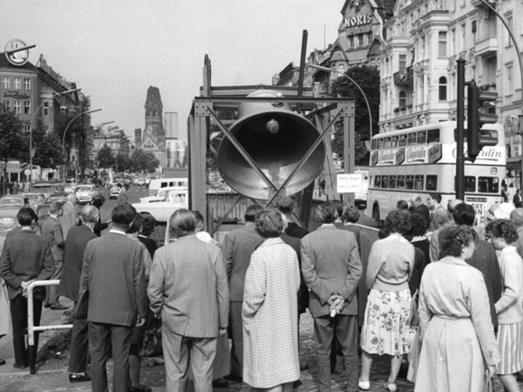In der Bildmitte läutet die riesige Glocke, mit dem Rücken zur Kamera stehen Schaulustige davor. Im Bildhintergrund steht die Kaiser-Wilhelm-Gedächtniskirche.