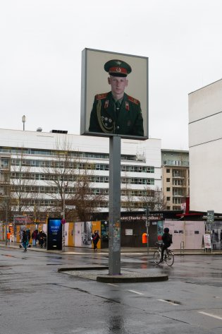 Checkpoint Charlie an einem regnerischen Tag, mittig auf einer Verkehrsinsel ein Leuchtkasten in einigen Metern Höhe, zeigt überlebensgroßes Foto eines russischen Soldaten, im Hintergrund Straßenkreuzung und Checkpoint Charlie BlackBox.
