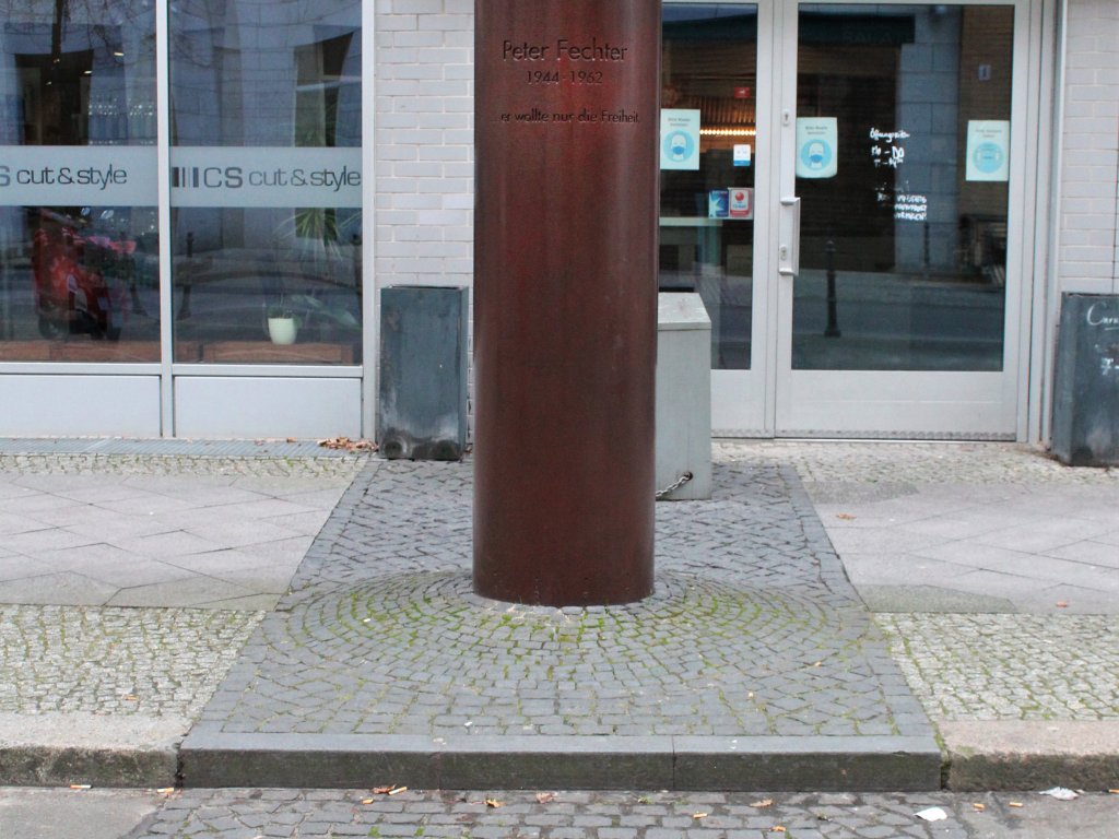 The bronze-coloured memorial column for Peter Fechter. In front of it is a line of bricks representing the path of the former Berlin Wall and a round granite plate.