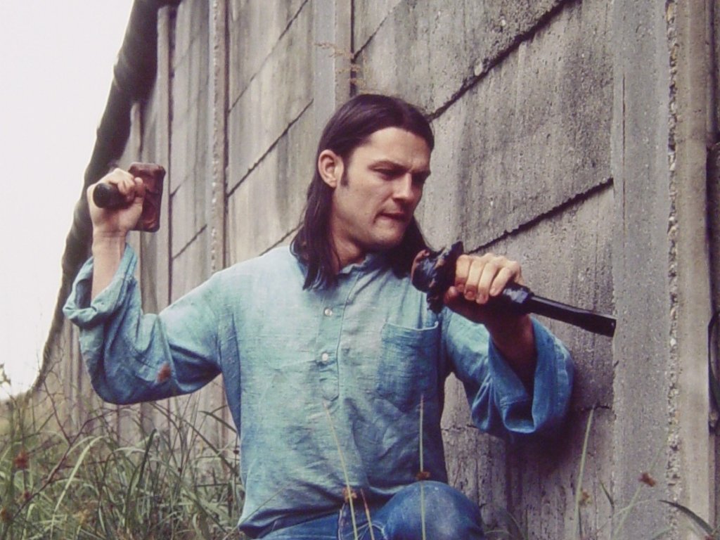 Stephan Elsner kniet im Gras neben der Mauer, die er mit Brecheisen und und Holzhammer bearbeitet.