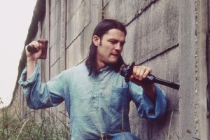 Stephan Elsner kniet im Gras neben der Mauer, die er mit Brecheisen und und Holzhammer bearbeitet.