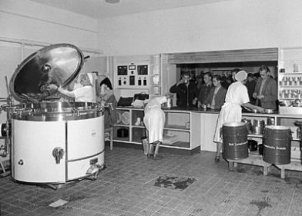 Der Blick ist vom inneren einer Großküche auf die Essensausgabe gerichtet. Drei Kantinenarbeiterinnen in weißen Kitteln bedienen die dicht an dicht stehenden Menschen auf der anderen Seite der Theke.