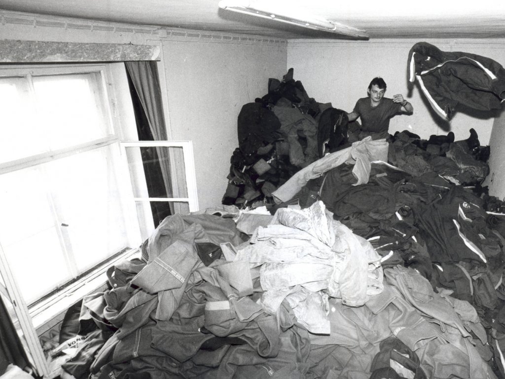 Ein Mann sitzt in einem Zimmer auf einem Berg von Uniformen und wirft von hinten eine Jacke durch den Raum.