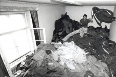 Ein Mann sitzt in einem Zimmer auf einem Berg von Uniformen und wirft von hinten eine Jacke durch den Raum.