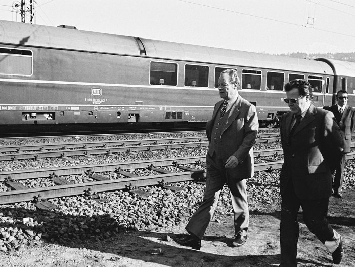 Willy Brandt und Günter Guillaume gehen entlang einer Bahnstrecke von rechts durchs Bild. Im Hintergrund steht ein Zug der Deutschen Bahn.