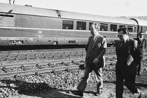 Willy Brandt und Günter Guillaume gehen entlang einer Bahnstrecke von rechts durchs Bild. Im Hintergrund steht ein Zug der Deutschen Bahn.