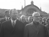Willy Brandt links und Willi Stoph rechts schauen seitlich an der Kamera vorbei. Hinter ihnen steht eine Menschenmenge. Im Hintergrund ist der Erfurter Hauptbahnhof zu sehen.