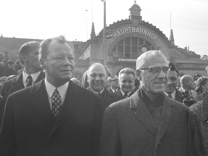Willy Brandt links und Willi Stoph rechts schauen seitlich an der Kamera vorbei. Hinter ihnen steht eine Menschenmenge. Im Hintergrund ist der Erfurter Hauptbahnhof zu sehen.