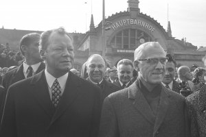 Willy Brandt links und Willi Stoph rechts schauen seitlich an der Kamera vorbei. Hinter ihnen steht eine Menschenmenge. Im Hintergrund ist der Erfurter Hauptbahnhof zu sehen.