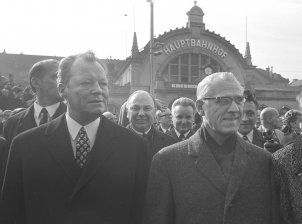 Willy Brandt links und Willi Stoph rechts schauen seitlich an der Kamera vorbei. Hinter ihnen steht eine Menschenmenge. Im Hintergrund ist der Erfurter Hauptbahnhof zu sehen.