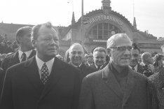 Willy Brandt links und Willi Stoph rechts schauen seitlich an der Kamera vorbei. Hinter ihnen steht eine Menschenmenge. Im Hintergrund ist der Erfurter Hauptbahnhof zu sehen.