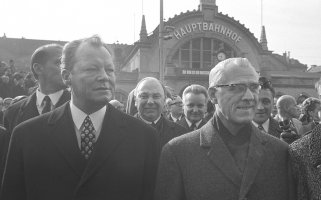Willy Brandt links und Willi Stoph rechts schauen seitlich an der Kamera vorbei. Hinter ihnen steht eine Menschenmenge. Im Hintergrund ist der Erfurter Hauptbahnhof zu sehen.