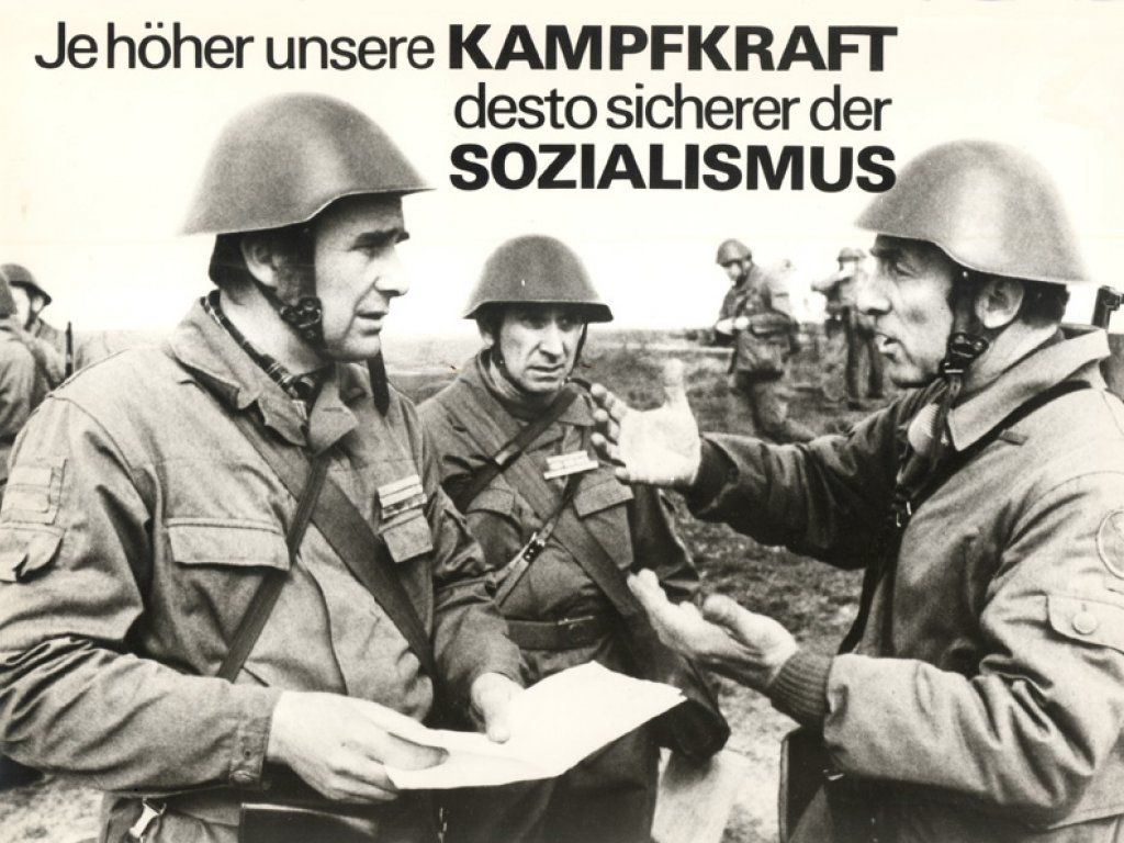 Drei uniformierte, bewaffnete Männer in Stahlhelmen diskutieren im Vordergrund. Im Hintergrund sind weitere Soldaten zu sehen. Das Bild ist beschriftet mit: Ja höher unsere Kampfkraft desto sicherer der Sozialismus.
