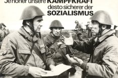 Drei uniformierte, bewaffnete Männer in Stahlhelmen diskutieren im Vordergrund. Im Hintergrund sind weitere Soldaten zu sehen. Das Bild ist beschriftet mit: Ja höher unsere Kampfkraft desto sicherer der Sozialismus.