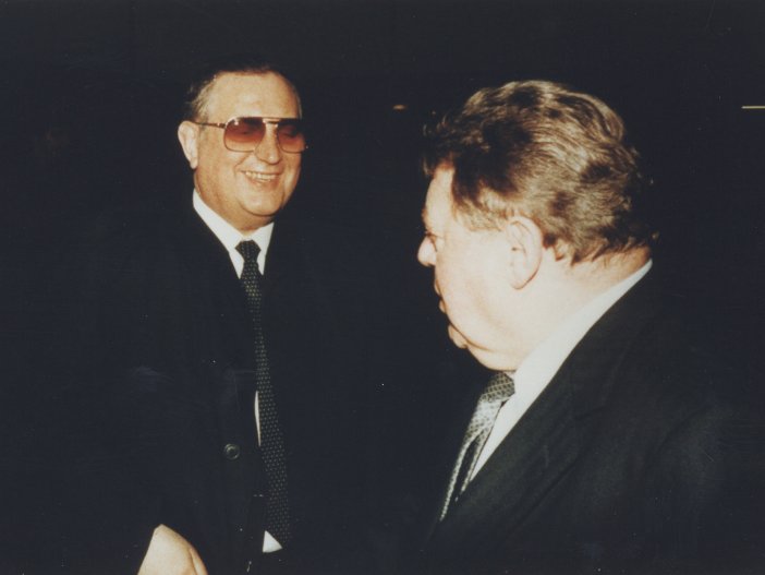 Schalck-Golodkowski und Franz Josef Strauß stehen einander zugewandt in der Dunkelheit, ihre Gesichter vom Blitzlicht erhellt. Schalck-Golodkowski ist frontal zu sehen, er trägt eine Sonnenbrille und grinst. Strauß ist abgewandt, man sieht ihn im Profil.