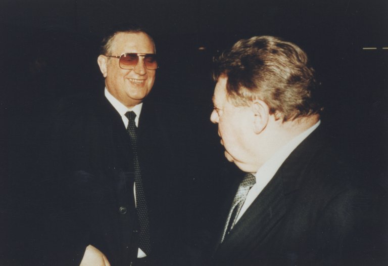 Schalck-Golodkowski und Franz Josef Strauß stehen einander zugewandt in der Dunkelheit, ihre Gesichter vom Blitzlicht erhellt. Schalck-Golodkowski ist frontal zu sehen, er trägt eine Sonnenbrille und grinst. Strauß ist abgewandt, man sieht ihn im Profil.