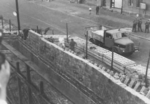 Unscharf im Vordergrund am linken Bildrand ist eine Hand mit Fotokamera zu erkennen. Quer durch das Bild zieht sich die Mauer aus Hohlblocksteinen, die gerade aufgebaut wird. Dahinter sieht man Arbeiter, eine Straßensperre aus Betonelementen, einen Lastwagen, drei Polizisten und zwei Passanten.