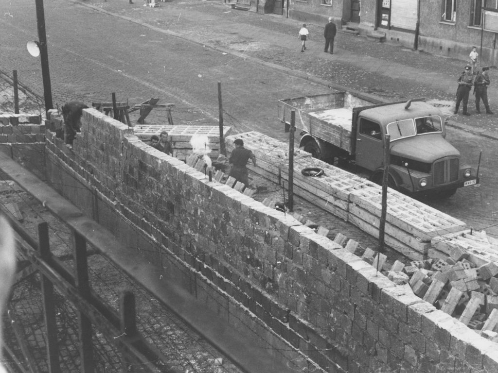 Tränengas gegen West-Berliner, die die Arbeiten zur Erhöhung der Mauer beobachten, 17. September 1961. Unscharf im Vordergrund am linken Bildrand ist eine Hand mit Fotokamera zu erkennen. Quer durch das Bild zieht sich die Mauer aus Hohlblocksteinen, die gerade aufgebaut wird. Dahinter sieht man Arbeiter, eine Straßensperre aus Betonelementen, einen Lastwagen, drei Polizisten und zwei Passanten.